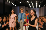 A Fashion Show of Young Designers with business women and men cheering for them, a young girl accepting an awrad trophy for winning the fashion show contest